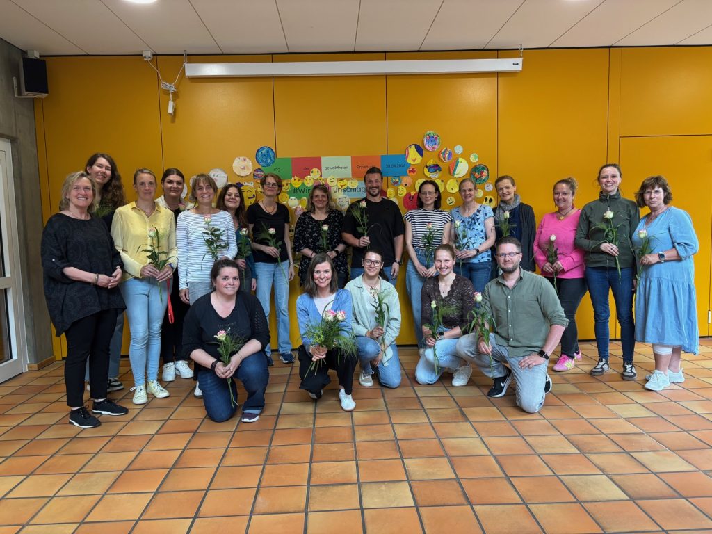 Lehrkräfte und Mitarbeitende der GGS Becke stehen mit Blumen vor einer gelben Wand und einem bunten Banner zum Tag der gewaltfreien Erziehung.